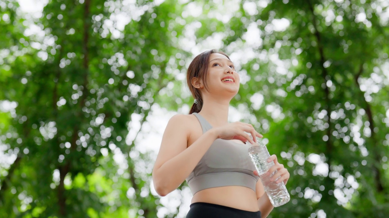 年轻的亚洲女子喝水从瓶中休息时，她的锻炼。快乐的青春期女性在公园里锻炼，休息休息，喝点水视频素材