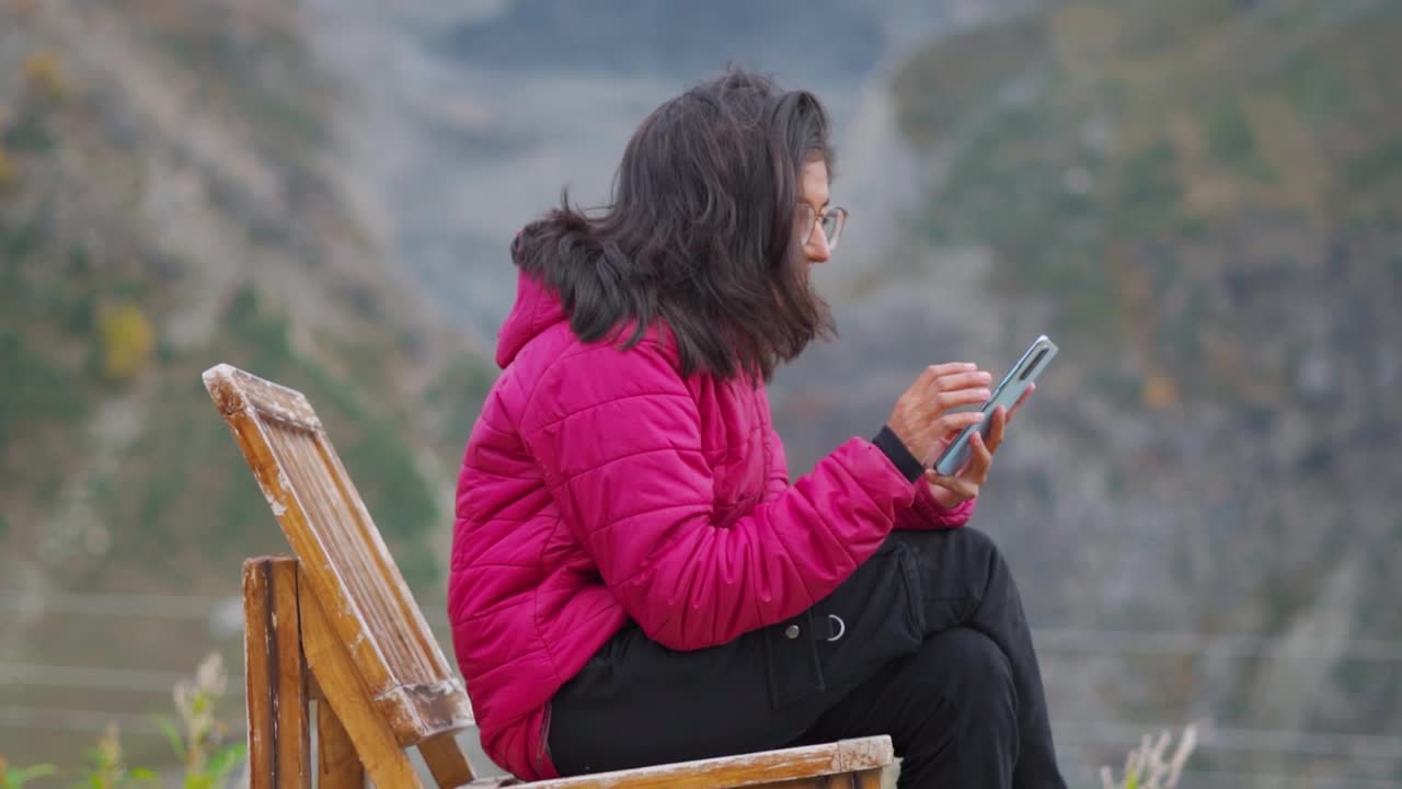 年轻的印度女子穿着冬季夹克，坐在椅子上使用手机的肖像。假日期间，女性在山的背景下放松。视频素材