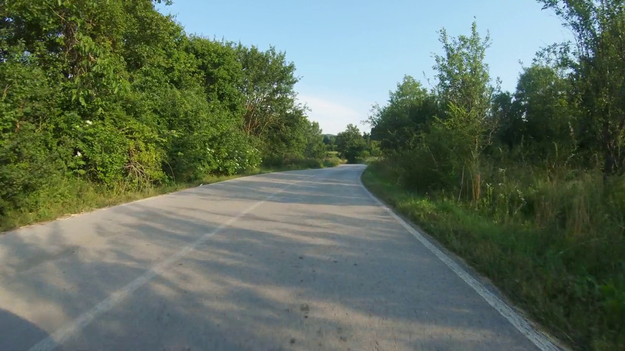 在树木环绕的蜿蜒起伏的乡间道路上缓慢行驶的汽车或摩托车。蓝天，夏日时光。观点拍摄视频素材