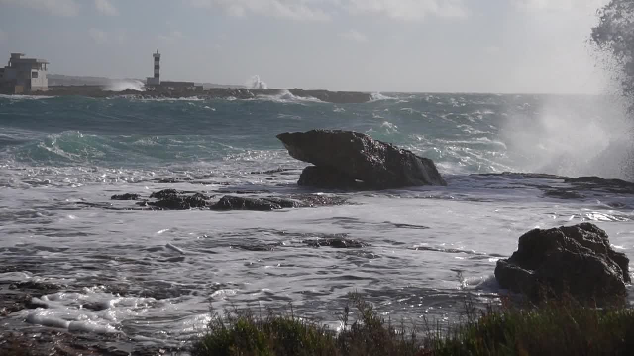 在暴风雨后的一个阳光灿烂的日子里，以灯塔为背景的海浪粉碎海岸线岩石的特写镜头视频下载