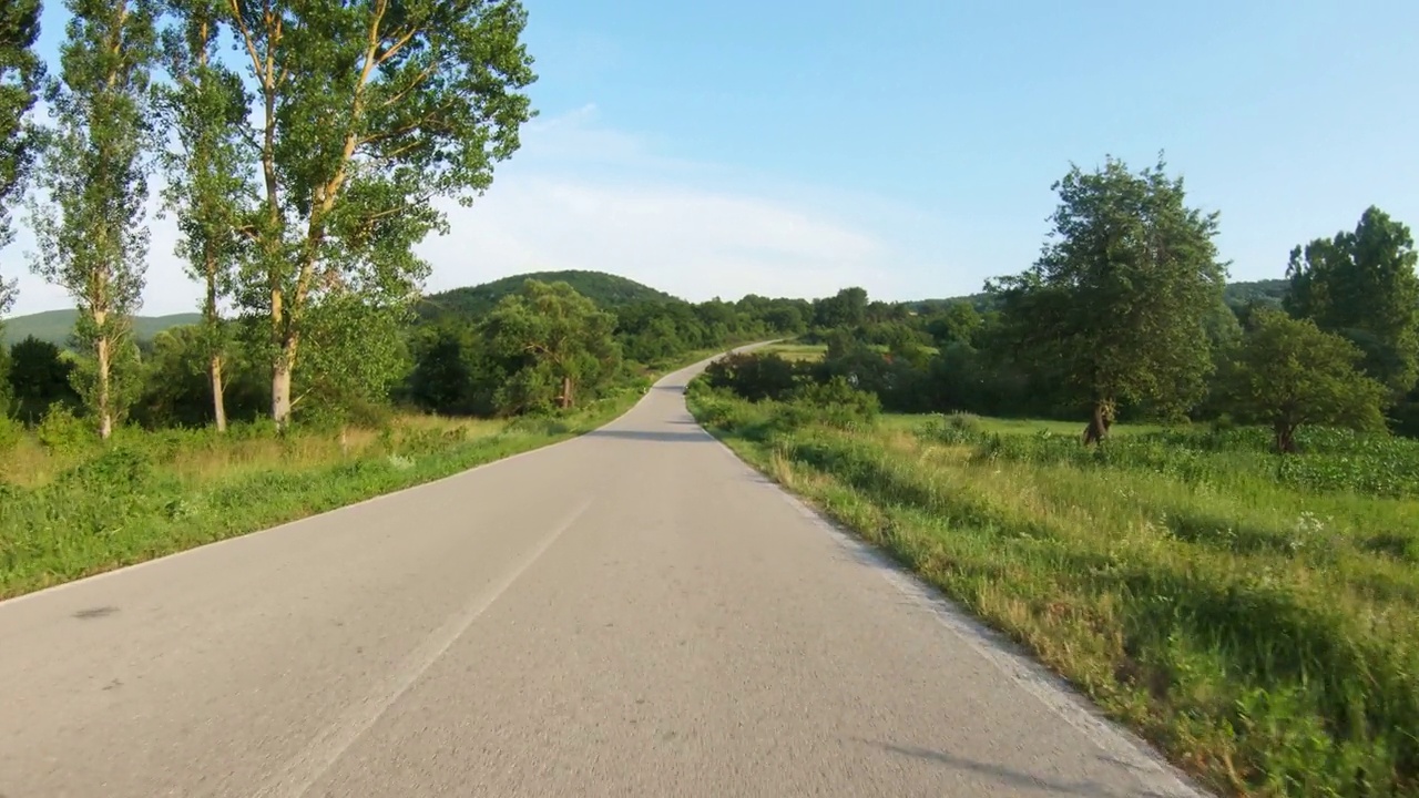 在被草地和树木环绕的乡间道路上缓慢地骑汽车或摩托车上山。蓝天，夏日时光。观点拍摄视频素材