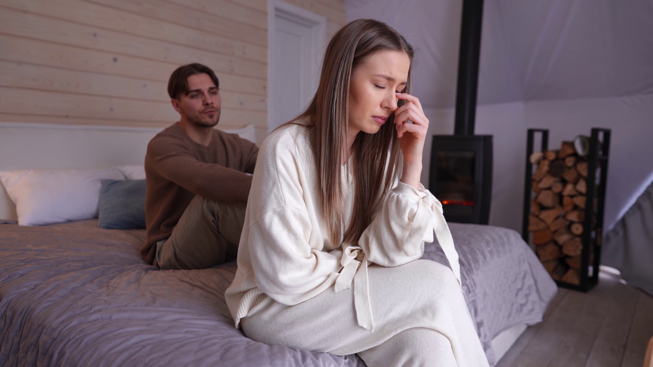 年轻美丽的女人哭着坐在床上与模糊的男人说话在背景借口背叛。被冒犯的心碎的白人妻子和有罪的丈夫在卧室室内的肖像。视频素材