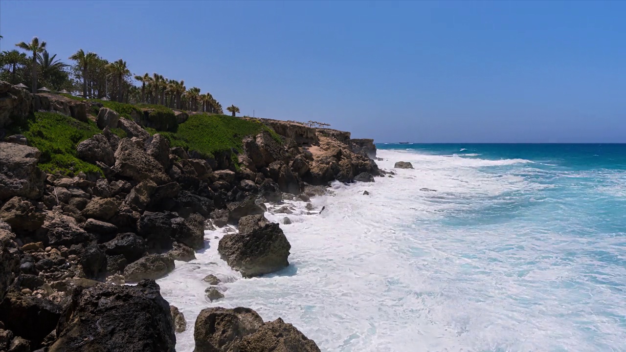 大海浪拍打着热带岛屿的岩石，背景是棕榈树和蓝色的天空和远处的船只视频素材