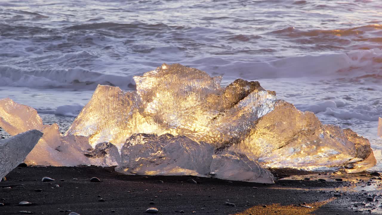 日落时冰山在黑色火山沙上闪闪发光，被海浪撞击的清晰冰块，冰岛著名的钻石海滩，8K分辨率拍摄视频素材