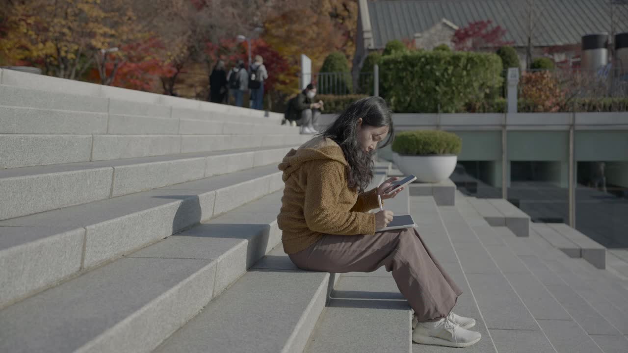 年轻的亚洲女性坐在台阶上，在城市里使用数字平板电脑。视频素材