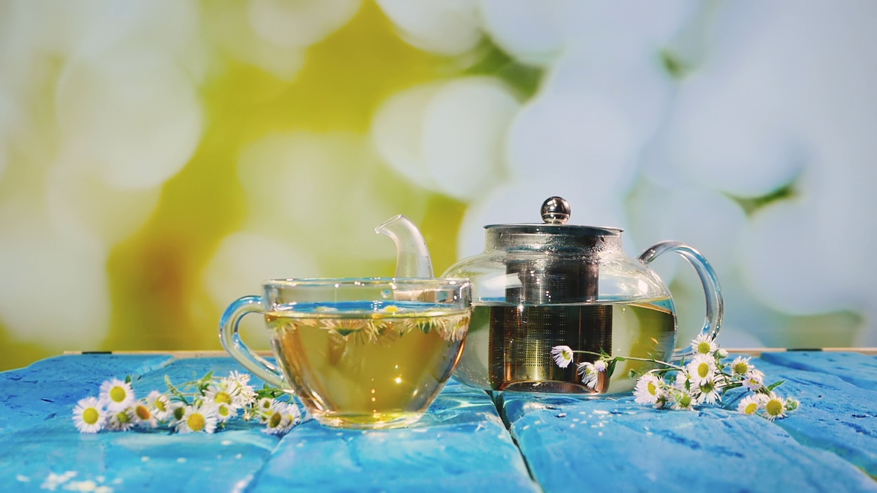 洋甘菊茶，茶杯和茶壶放在蓝色的木桌子上。芬芳的药茶与花瓣在夏天的背景。健康饮料。物理疗法输液视频素材