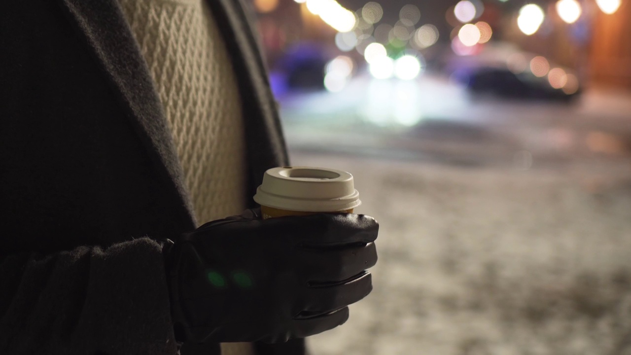 特写一名不认识的男子的手，他站在冬天积雪的街道上，用纸杯在户外享受咖啡，背景是行驶的汽车视频素材