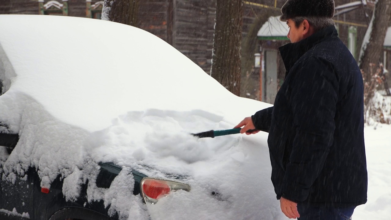 一个人用一把干净的刷子清理汽车引擎盖上的雪，正在下雪。视频素材