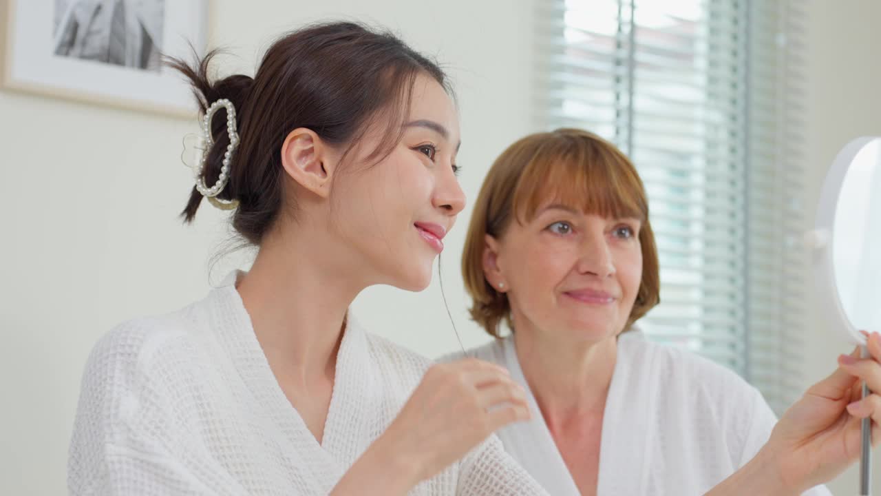 亚洲妇女在和母亲擦完乳液后看着镜子。迷人的年轻女孩和年长的妈妈穿着浴袍坐在床上，感到幸福和自豪的健康美容和护肤。视频素材