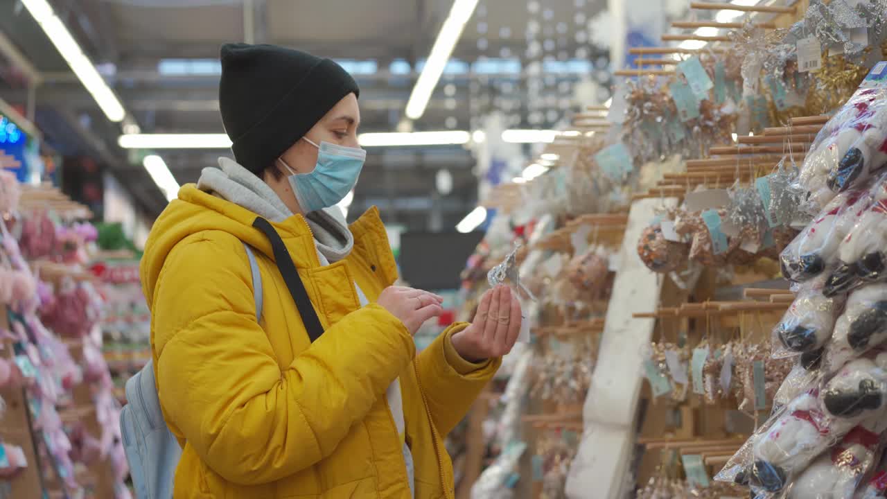 在购物中心的圣诞市场上，身穿黄色冬季夹克的妇女正在挑选圣诞装饰品。成年白人女性在购买医用防护口罩的新年装饰品视频素材