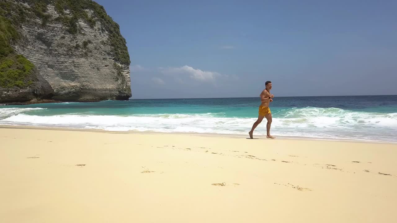 年轻快乐的运动旅行者沿着绿松石水和白色泡沫的波浪在热带海滩的沙滩上奔跑。印度尼西亚努沙佩尼达的克林金海滩。旅游、休闲、度假视频下载