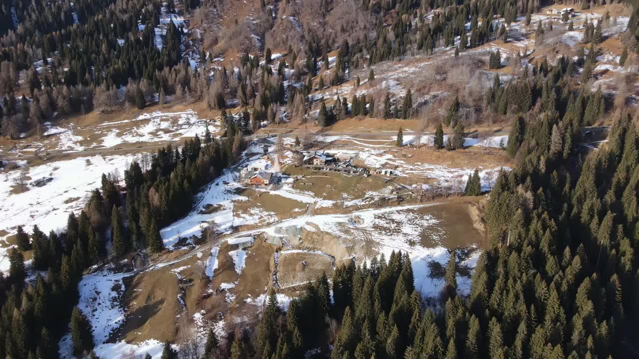 鸟瞰阳光下美丽的松树森林全景，巍峨的瑞士阿尔卑斯山脉覆盖着白雪，森林中心的木屋、小木屋和房屋为游客提供目的地，瑞士冬季休闲度假胜地视频素材