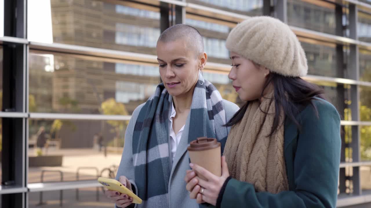 开朗的商务女性走路和使用手机。年轻的亚洲女性和中年白种人剃了头发的女士在办公楼附近讨论工作视频素材