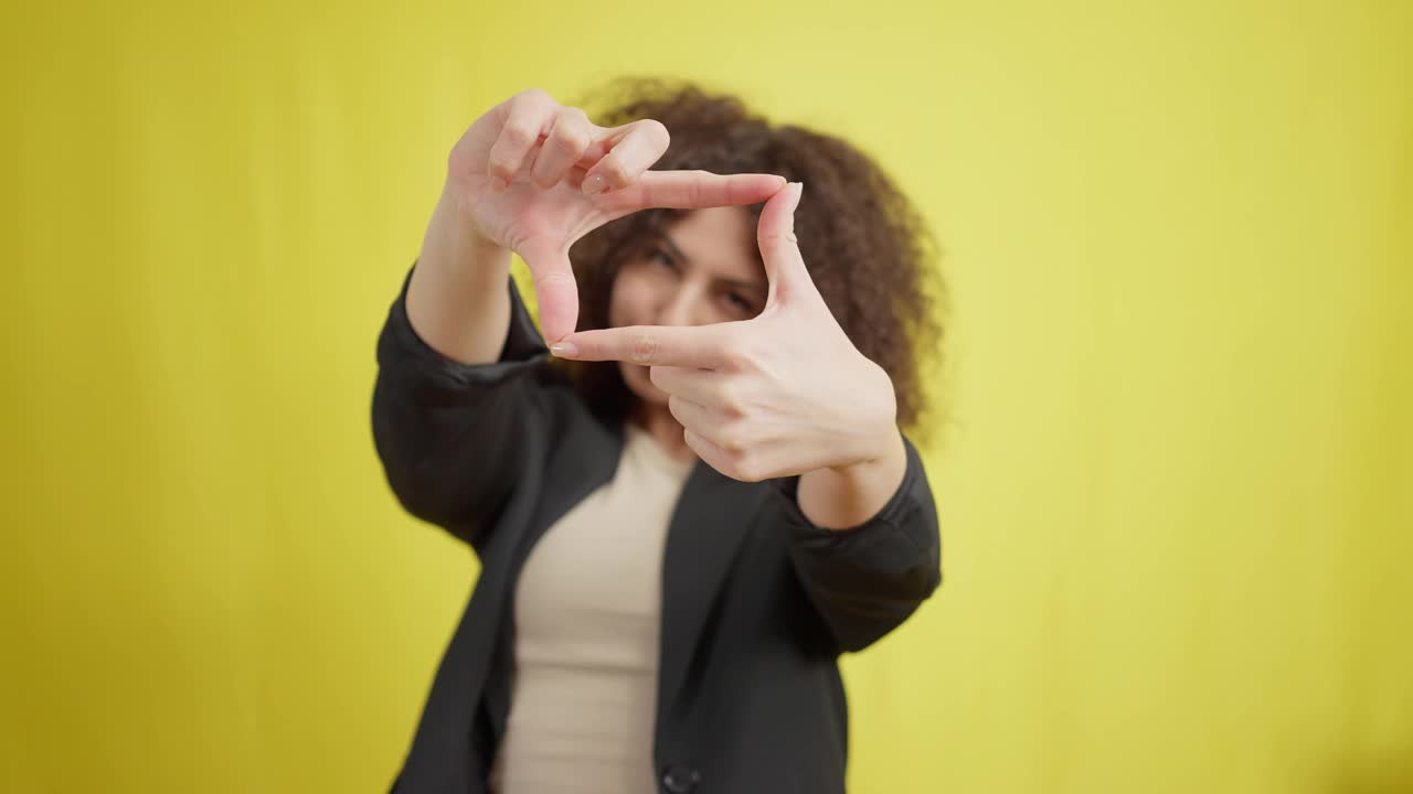 特写女性手方形模仿相机和模糊的年轻女子站在黄色背景。正面观点积极自信的高加索社交媒体视频博主摆姿势拍摄博客。视频素材