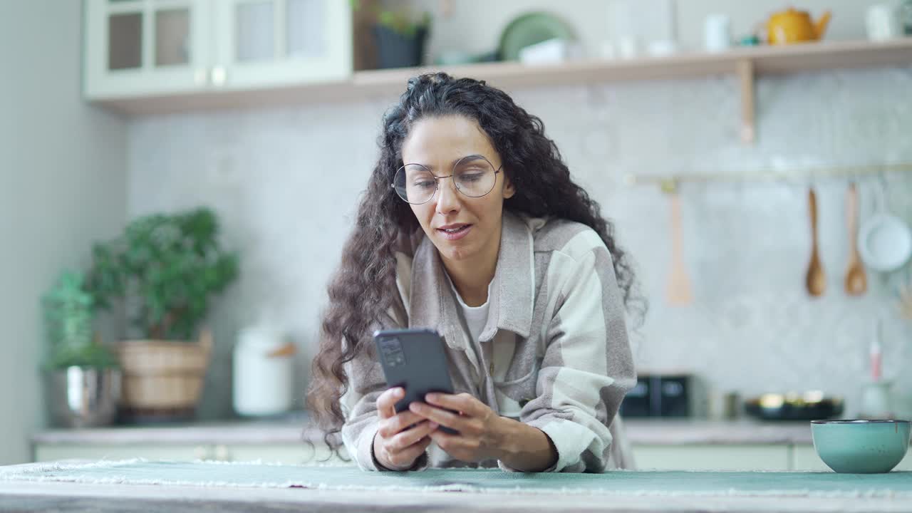 惊讶的女人戴眼镜非常高兴后阅读短信在手机上在家里兴奋卷发黑发看着智能手机屏幕视频素材