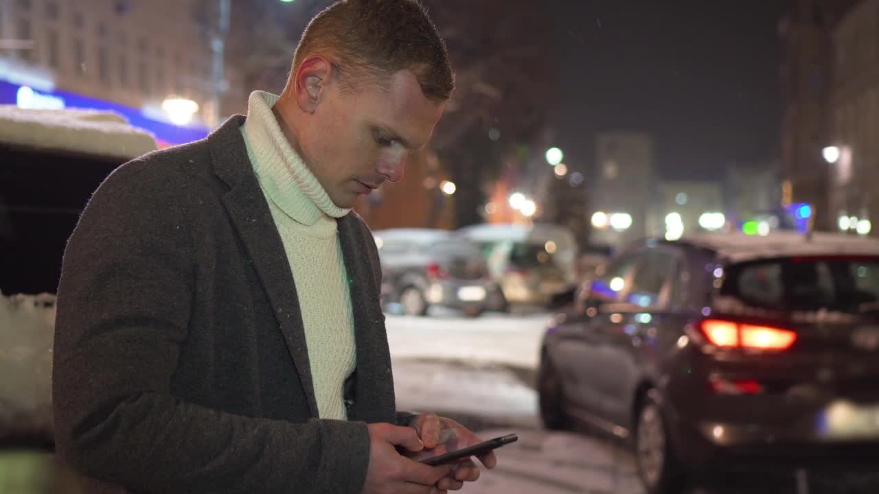 在下雪的夜晚，在拥挤的户外街道上，一辆车路过，一名微笑的帅哥一边看着手机屏幕一边使用智能手机视频素材