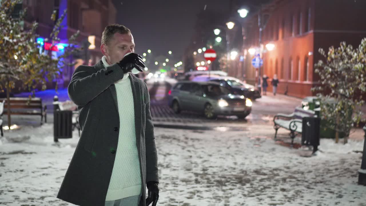 英俊的男子在户外享受纸杯咖啡，站在冬天积雪的街道上，背景是行驶的汽车视频素材