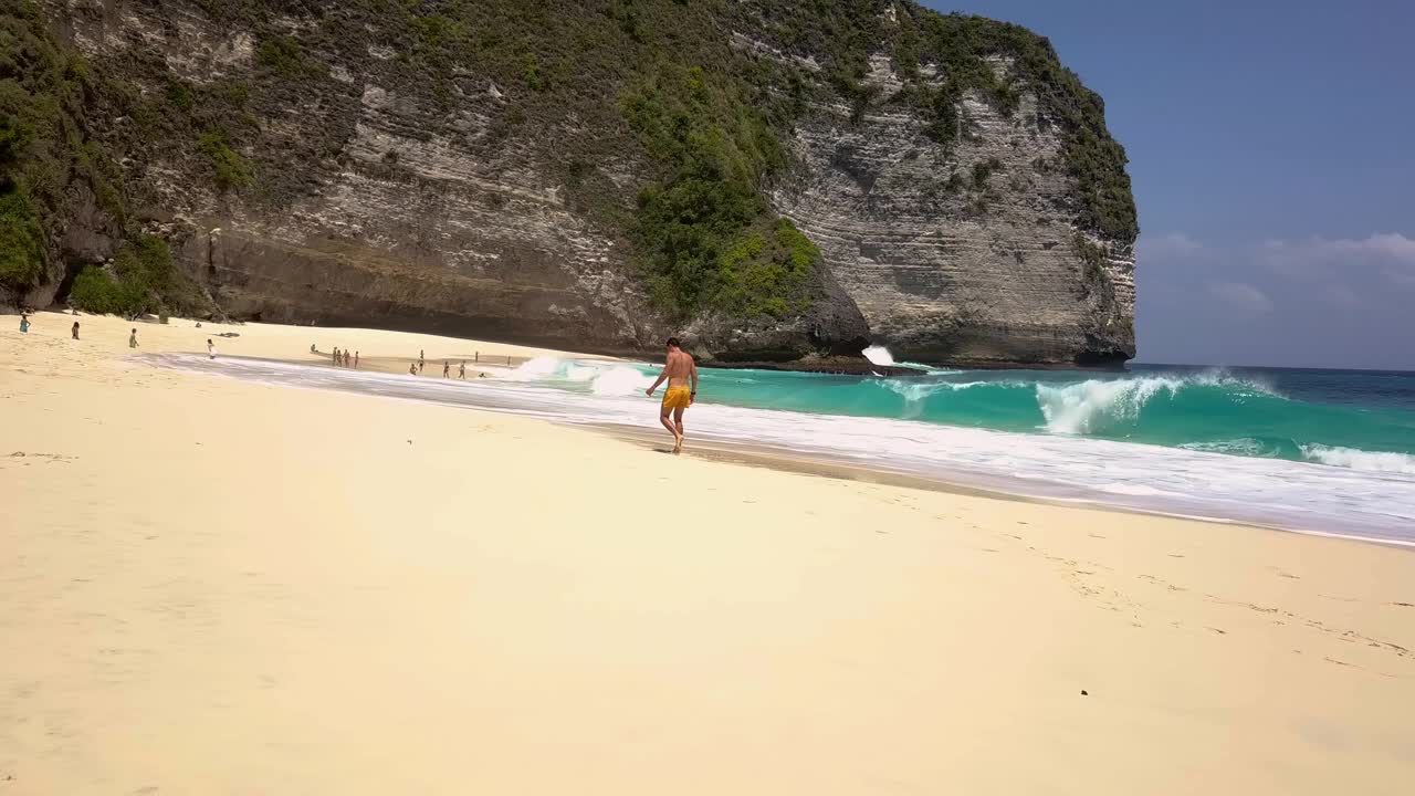 年轻快乐的运动旅行者沿着绿松石水和白色泡沫的波浪走在热带海滩的沙滩上。Kelingking海滩。旅游、度假。2022年8月25日，印度尼西亚努沙佩尼达视频素材