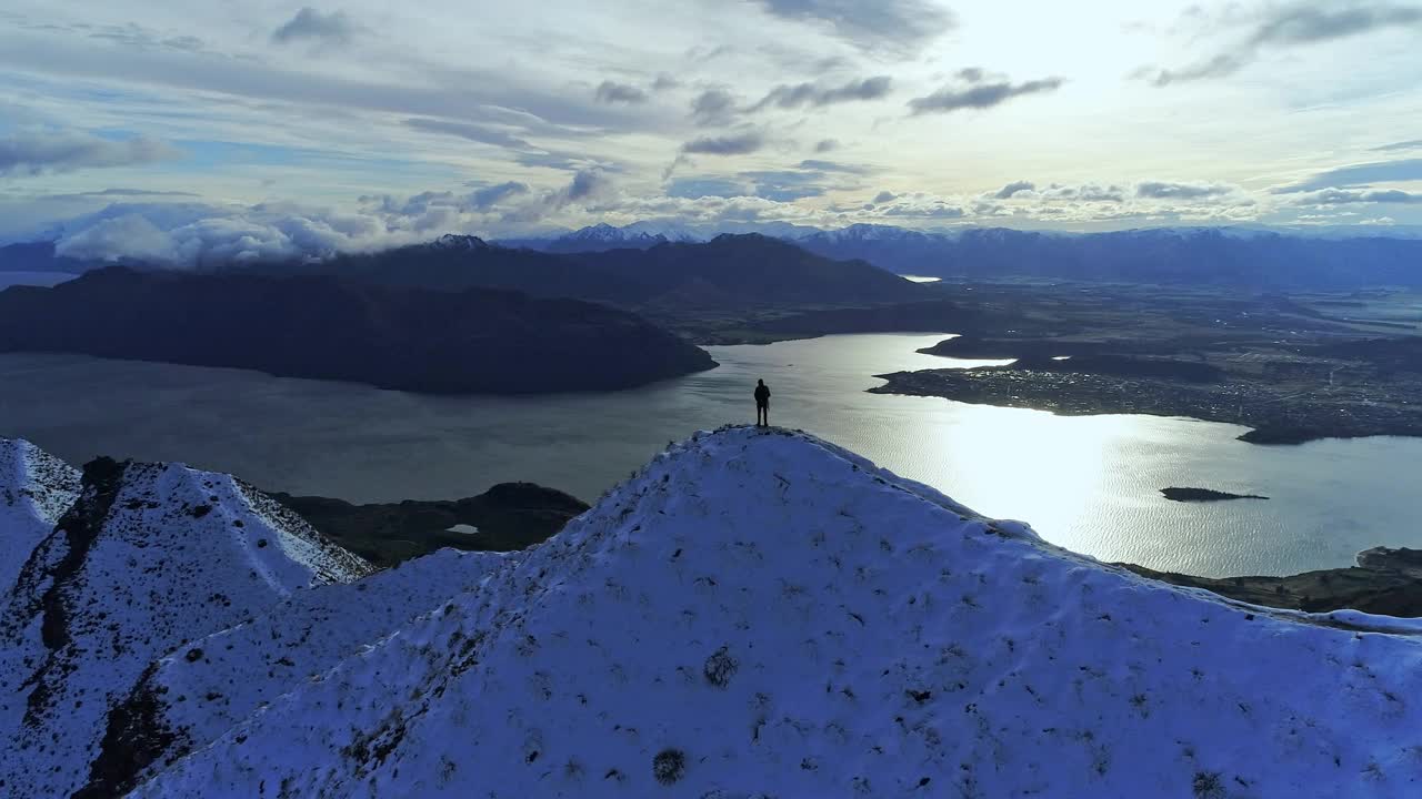 一名男性徒步旅行者站在山脊上，欣赏着雪山和周围大湖的壮丽自然风光视频素材