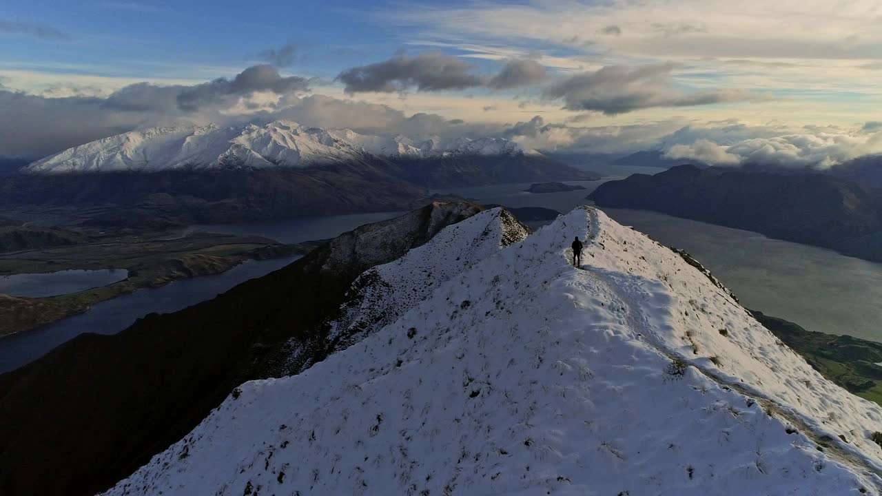 一名男性徒步旅行者站在山脊上，欣赏着雪山和周围大湖的壮丽自然风光视频素材