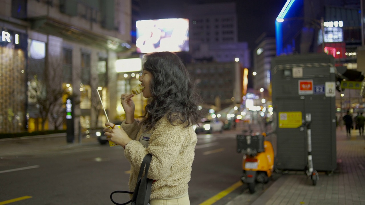 一名年轻的亚洲女子端着一碗汤，吃着挂在木棒上的鱼丸。，韩国视频素材