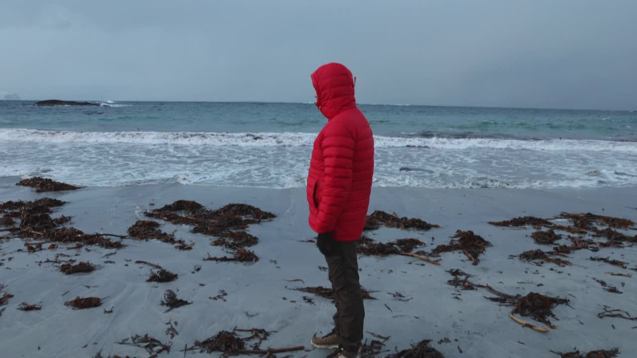 一名穿着红色夹克的男子在挪威风景优美的海滩上凝视着冬日的日落视频素材