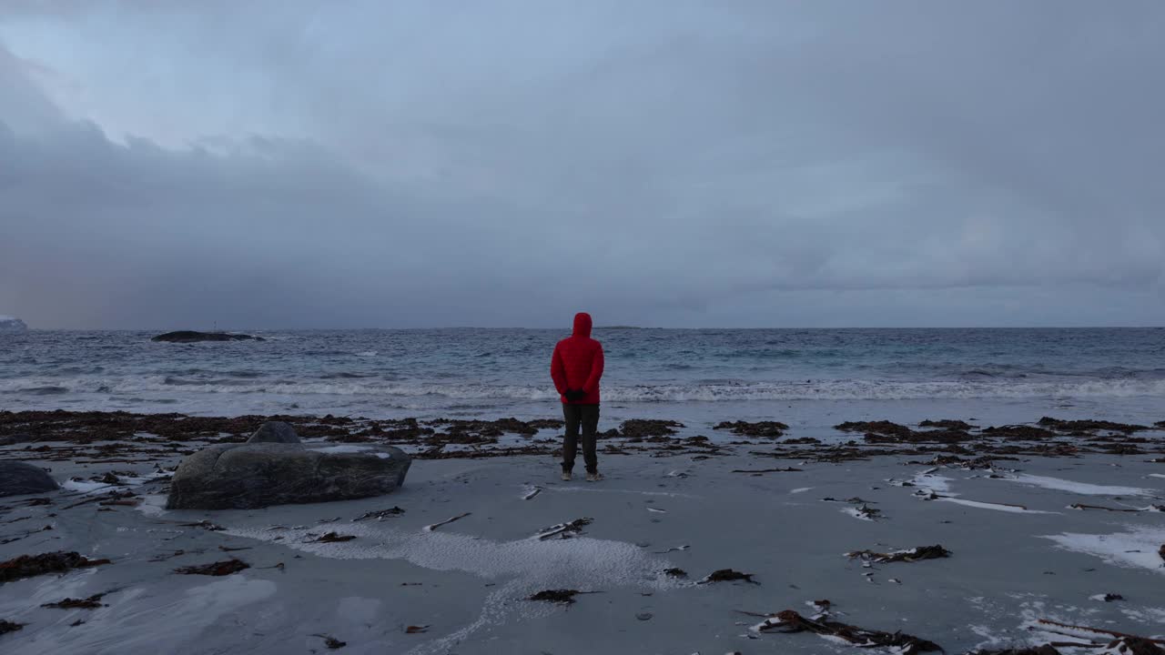 一名穿着红色夹克的男子在挪威风景优美的海滩上凝视着冬日的日落视频素材