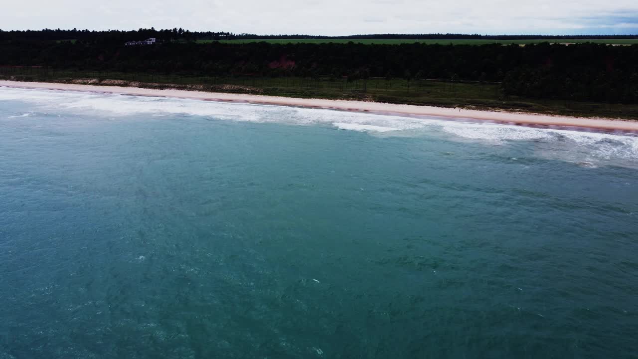 摄像机从深蓝色的海洋接近一个原始的巴西海滩，显示椰子树，红棕色的砾石路航拍视频视频素材