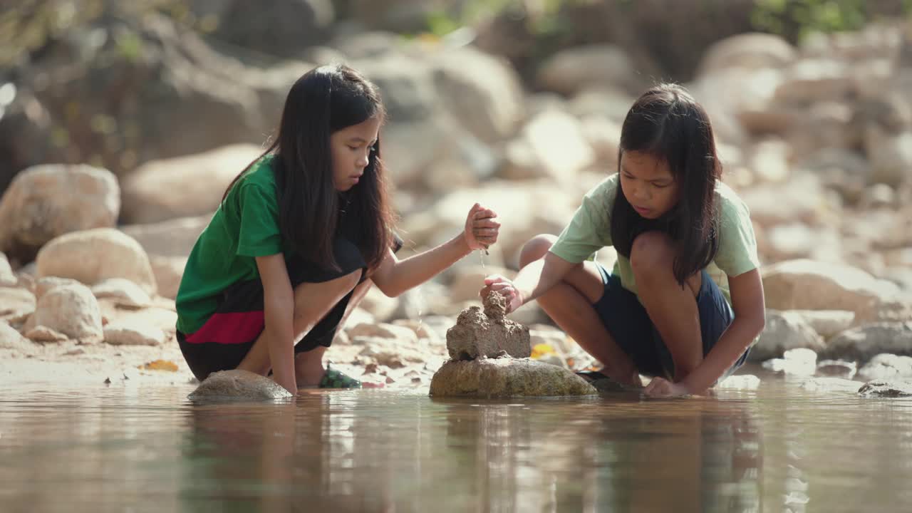 两个亚洲小女孩一起在河里玩沙子的慢动作镜头。孩子们正在玩沙玩水。孩子们在户外活动，享受大自然的假期。视频下载