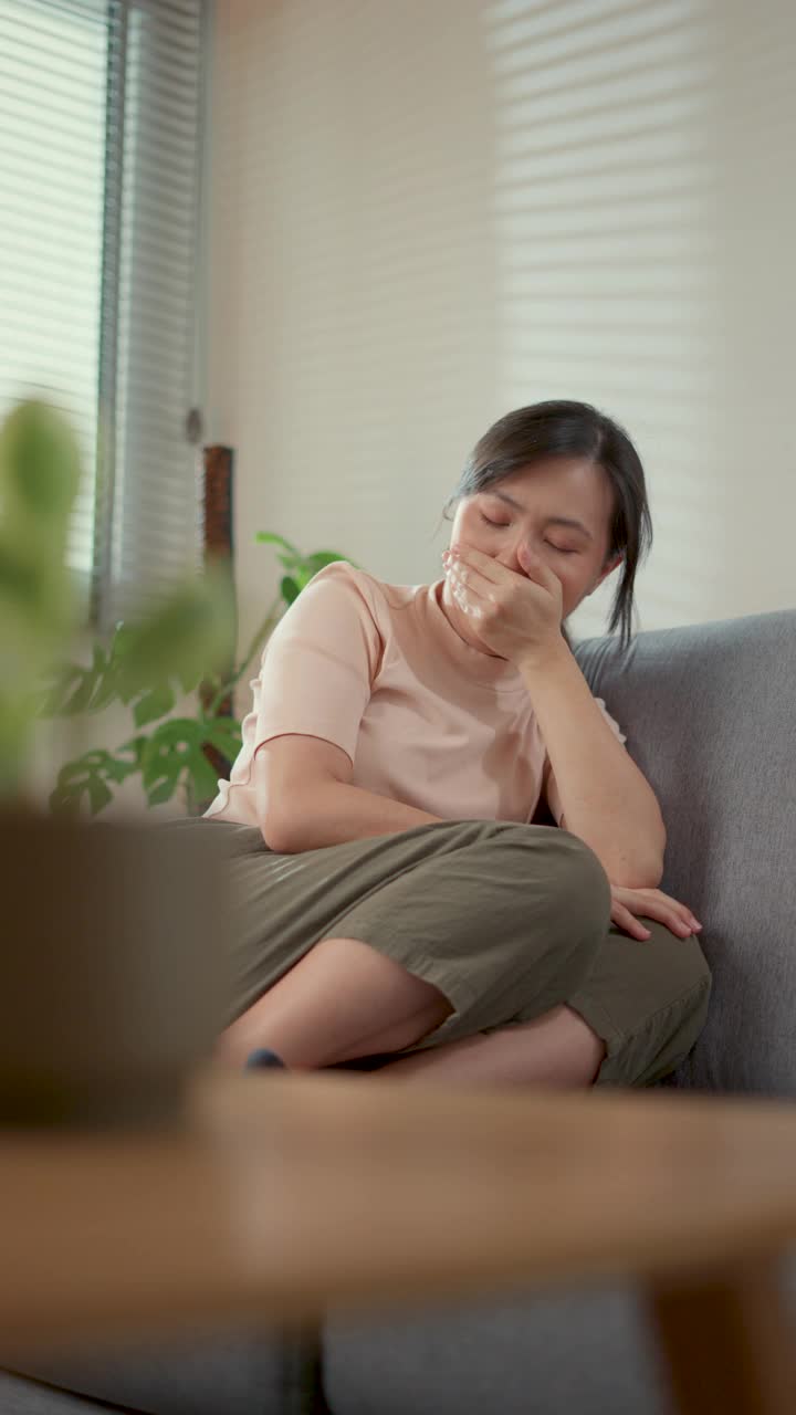 一名亚洲妇女坐在家里客厅的沙发上头痛。视频素材