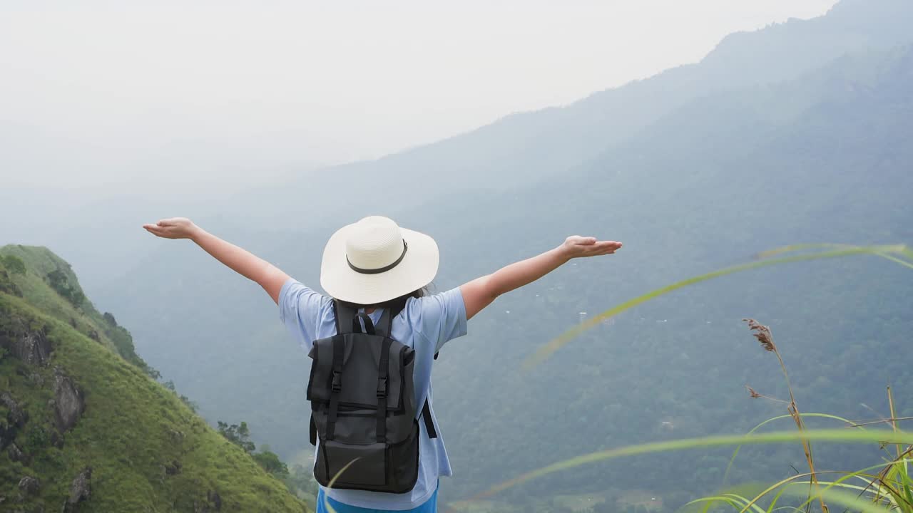 戴着帽子的女游客高举双臂站在山的尽头。视频素材