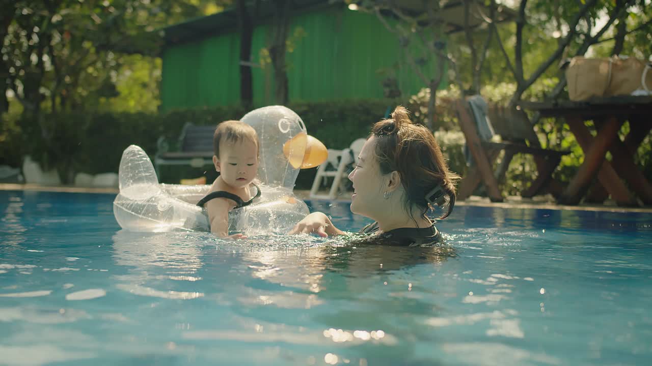 快乐的亚洲家庭妈妈和可爱的蹒跚学步的女儿一起在游泳池玩水，妈妈带着爱和微笑在假期的夏天抱着小孩视频素材