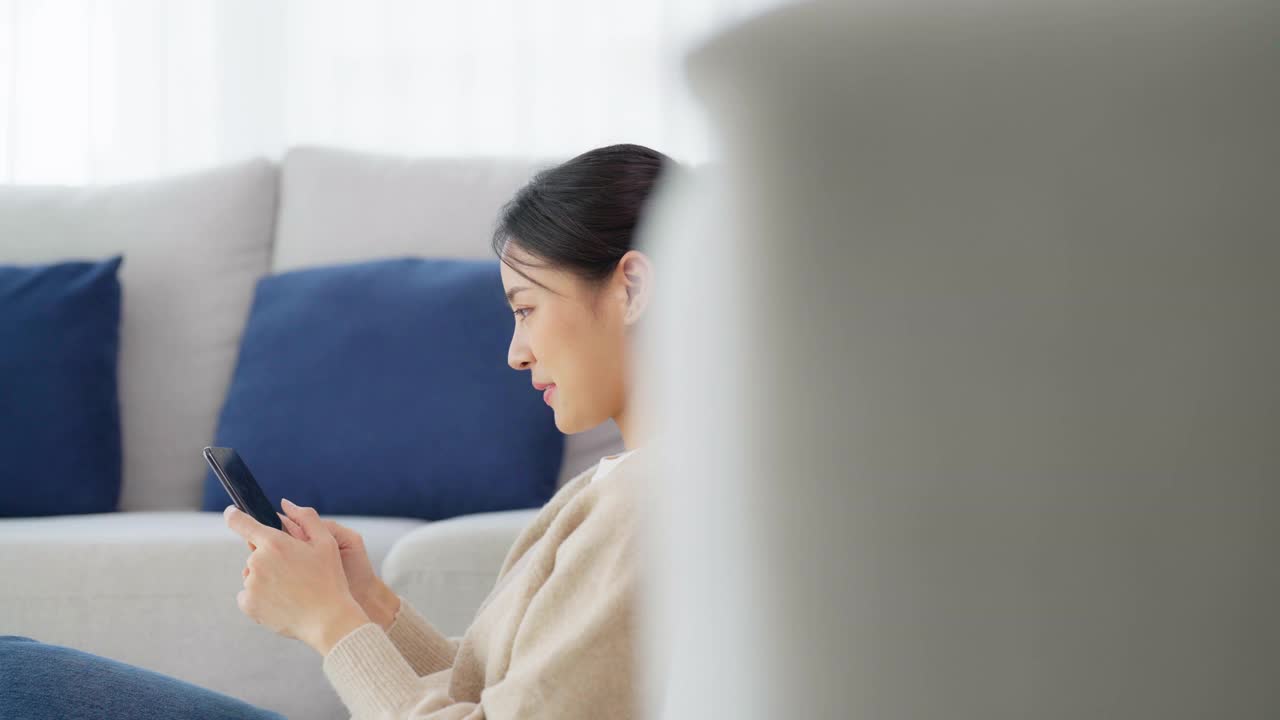 快乐的年轻亚洲女子在家里放松。女人微笑着坐在沙发旁，手里拿着手机。女孩用视频和朋友通话视频素材