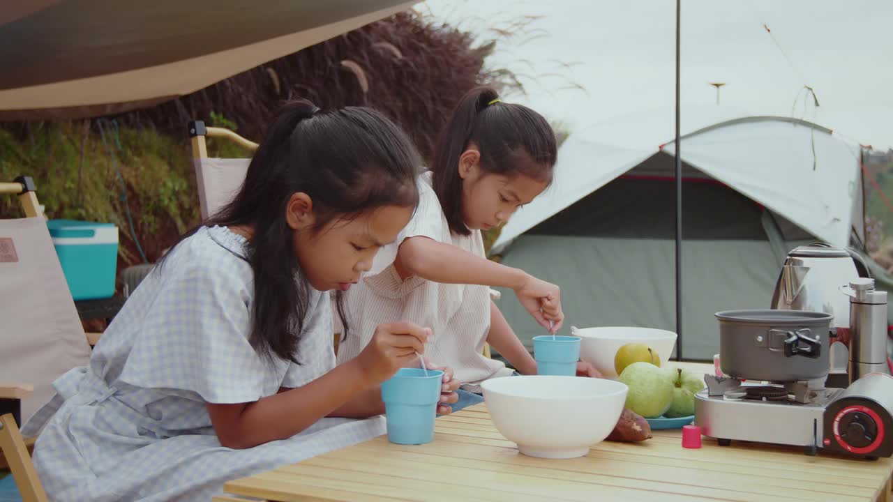 两个亚洲小女孩在和家人一起露营时吃早餐，一起享受美丽的大自然。孩子们享受大自然，在户外做有趣的活动。视频下载