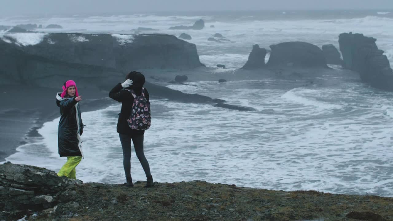 两个旅游女孩站在冰岛的悬崖边上，看着大海里的岩石和海浪。冬天的冰岛视频素材