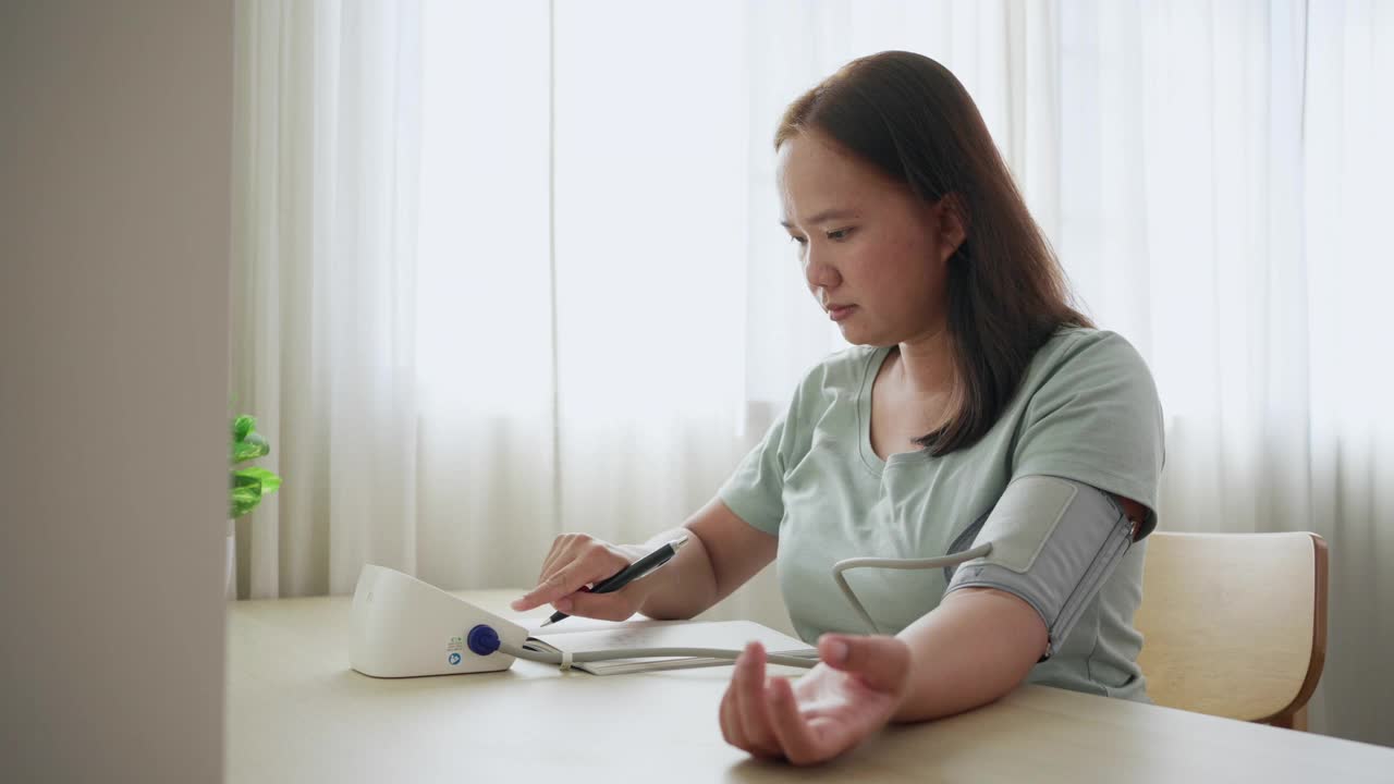 亚洲女性在家里的客厅里监测和检查血压并记录。常规自我医学检测视频素材