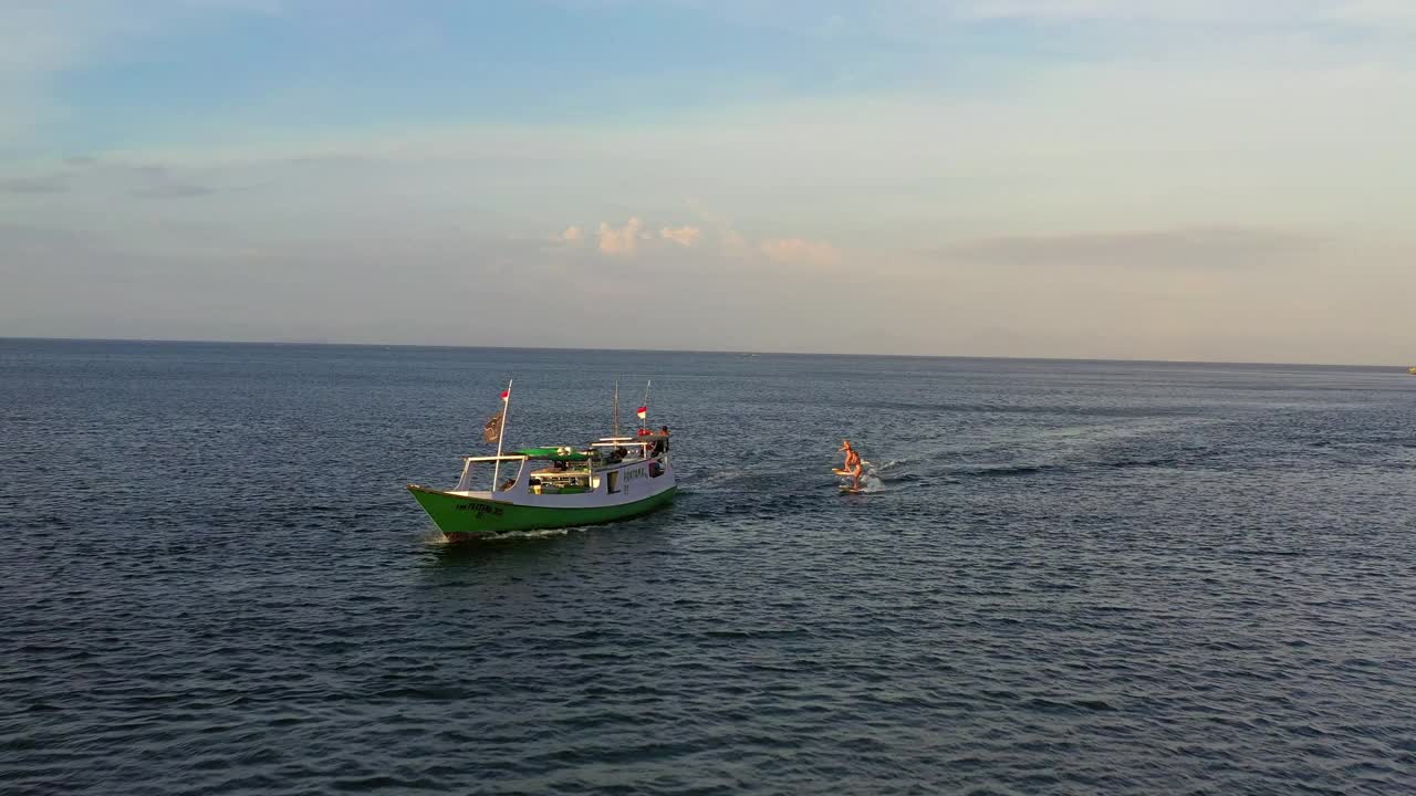 航拍的女性冲浪者在船后面冲浪，无人机在阳光灿烂的一天在海上向后飞行-龙目岛，印度尼西亚视频素材