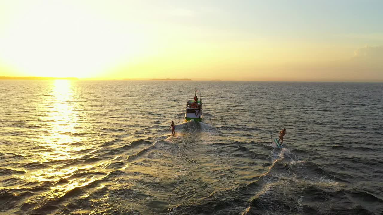 航拍女性朋友在海上船只后面冲浪，日落时无人机飞过海洋——印度尼西亚龙目岛视频素材