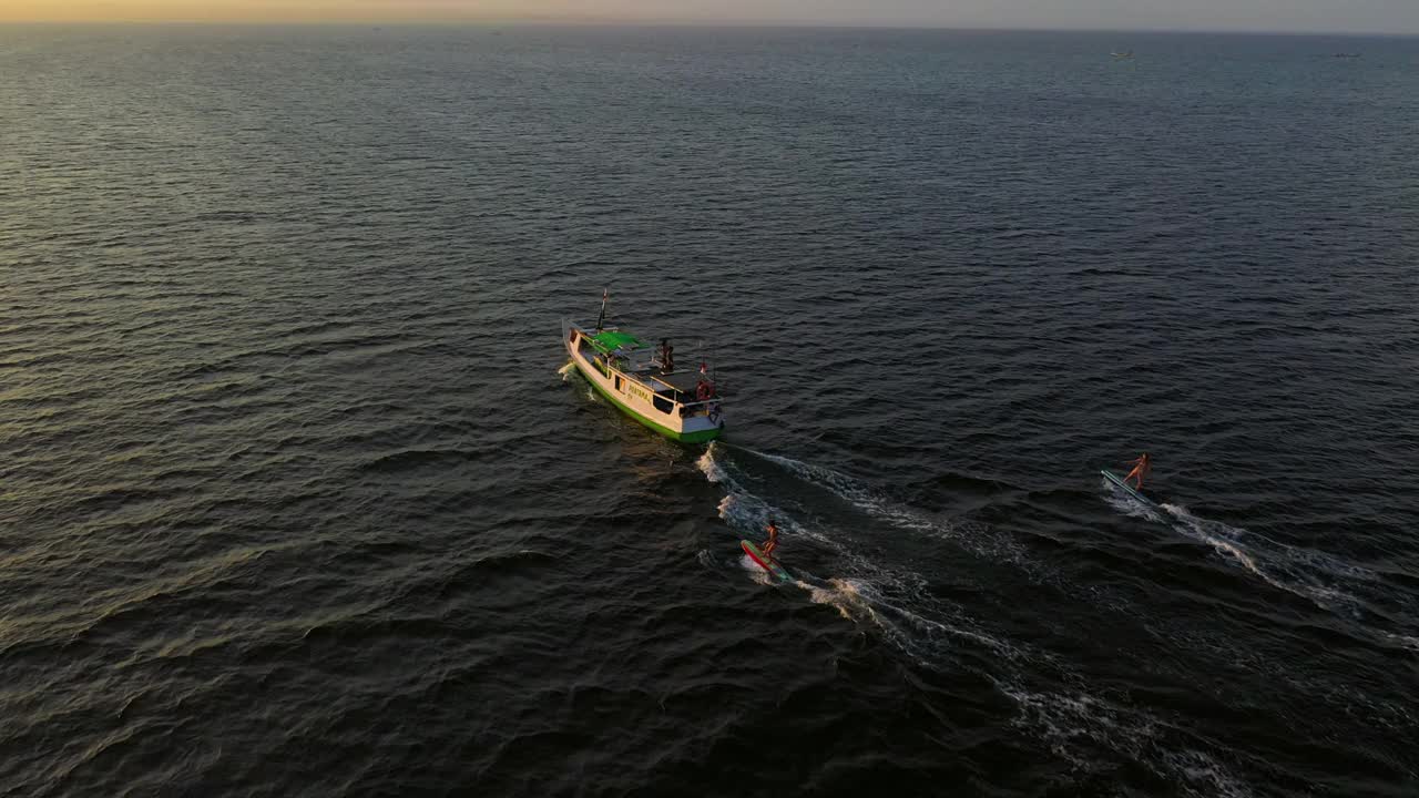 航拍拍摄冲浪者在海上移动的航海船只后面冲浪，日落时无人机向前飞行-龙目岛，印度尼西亚视频素材