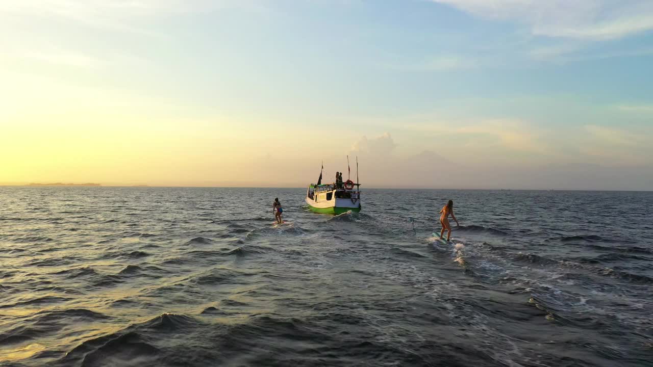 航拍冲浪者朋友在海上船只后面冲浪，无人机在日落期间在海上飞越海浪-龙目岛，印度尼西亚视频素材