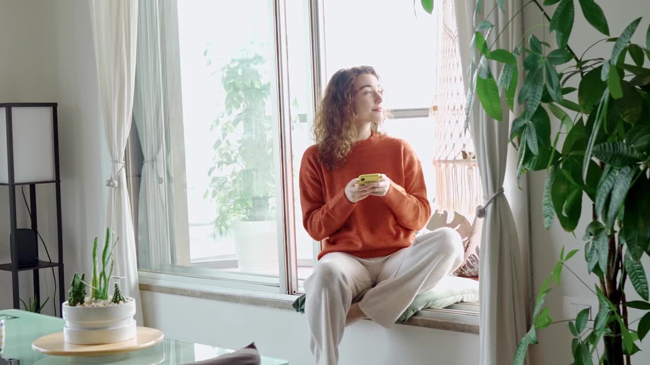 快乐的年轻女子使用手机坐在窗台上在家里。视频素材