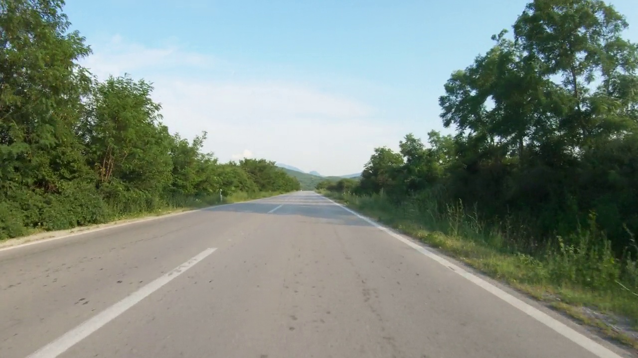 在树木环绕的乡间道路上缓慢行驶的汽车或摩托车。远处的群山。蓝天，夏日时光。观点拍摄视频下载