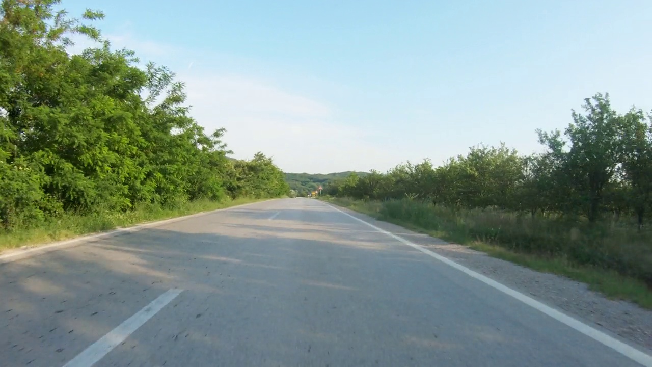 在被树木和草地环绕的崎岖的乡村道路上驾驶快速的汽车或摩托车。远处的群山。蓝天，夏日时光。观点拍摄视频下载