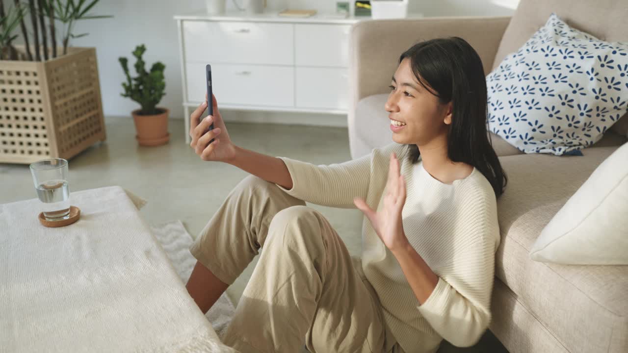 亚洲女性坐在地板上，拿着智能手机和朋友在手机屏幕上交流视频素材