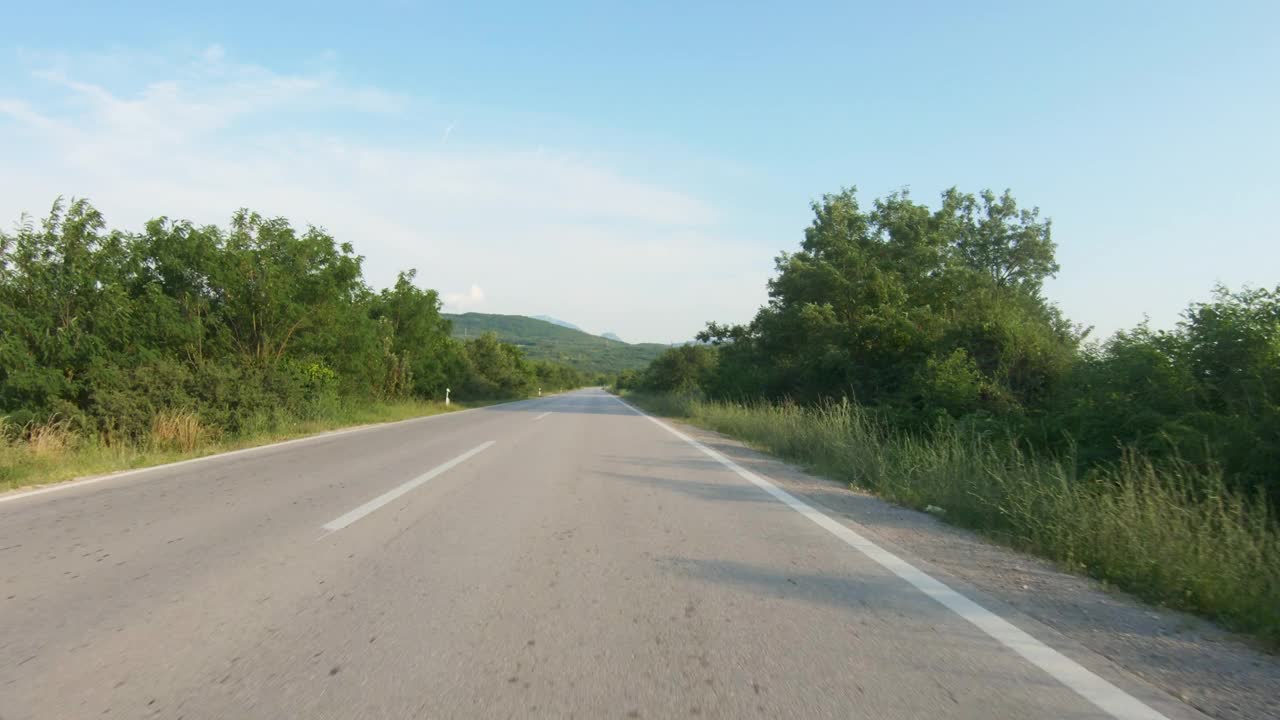 在树木环绕的笔直的乡村道路上缓慢地驾驶汽车或摩托车。远处的群山。蓝天，夏日时光。观点拍摄视频下载