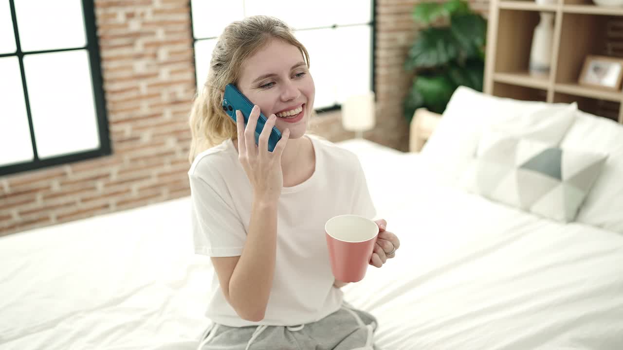 年轻的金发女子在卧室喝咖啡，用智能手机说话视频素材