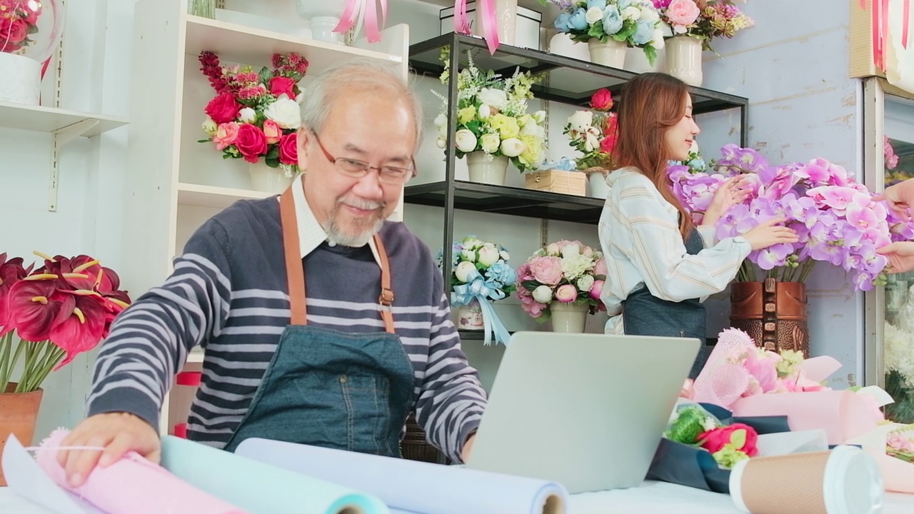 一位年长的亚洲男性老板在一家色彩缤纷的花店工作，手里拿着一台笔记本电脑。视频素材