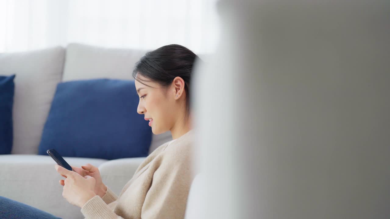 快乐的年轻亚洲女子在家里放松。女人微笑着坐在沙发旁，手里拿着手机。女孩用视频和朋友通话视频素材