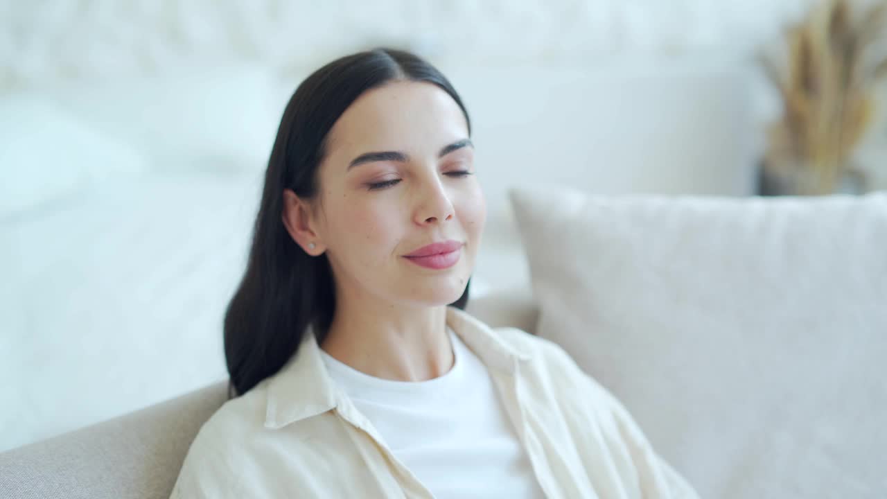 美丽的黑发年轻女士在舒适的沙发上放松，手在头后面在明亮的客厅。微笑平和的女孩独自在家享受平静的时光视频素材