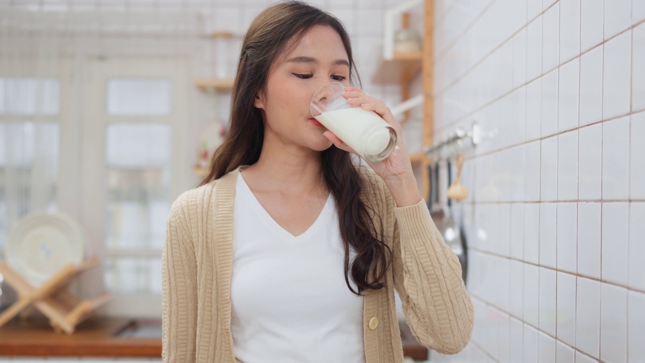 亚洲漂亮的女人拿着一杯牛奶站在家里的现代厨房微笑。早上喝牛奶提神。周末活动，健康理念。有机和减肥食品。视频素材